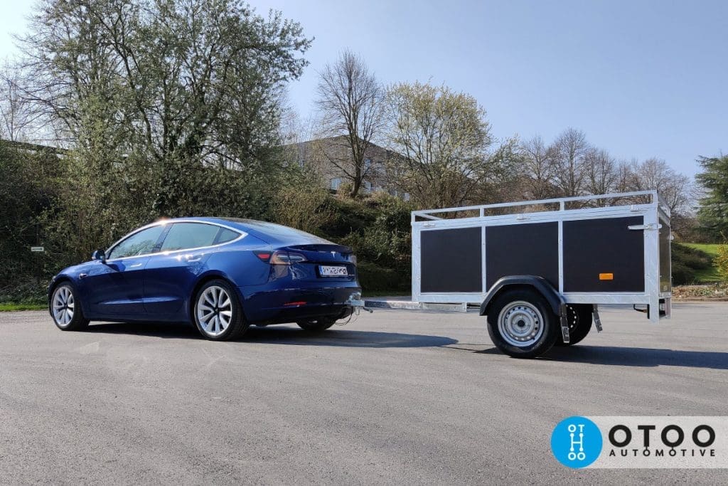 Een nieuwe ongeremde bagagewagen met enkele as die gekoppeld is aan de trekhaak van een blauwe elektrische Tesla auto in België bij mooi weer. De bovenzijde van de bagagewagen is voorzien van extra profielen om materieel op te plaatsen en te zekeren.